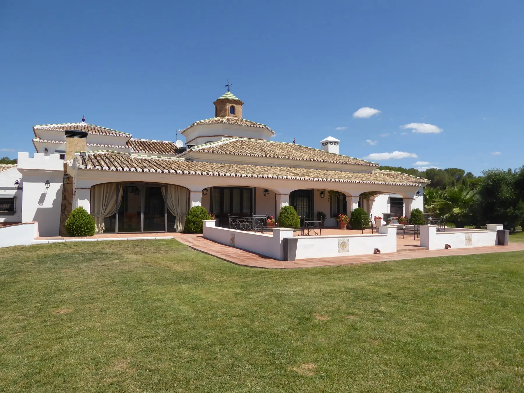 La casa principal vista desde el césped: el porche de arcos, la cúpula sobre el tejado de teja y los olivos al fondo, bajo un cielo azul