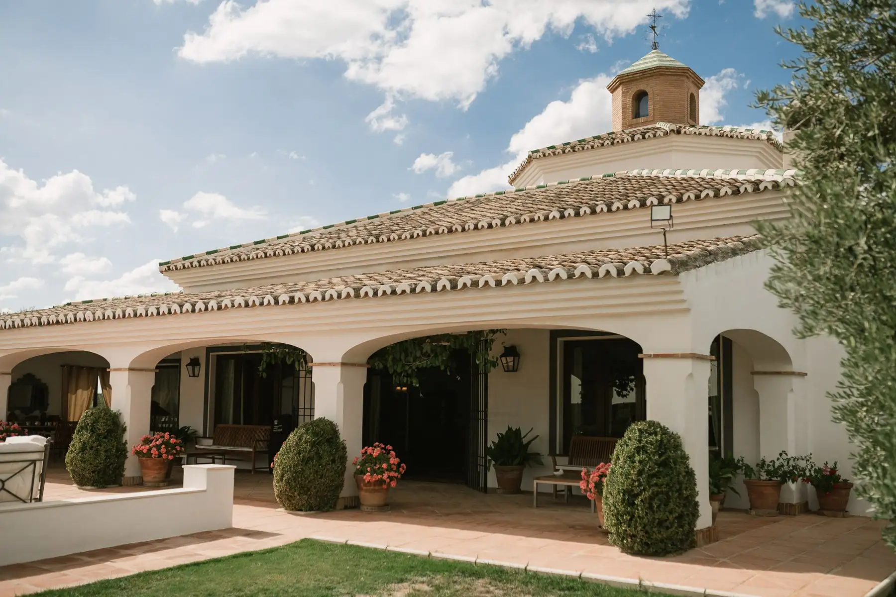 La casa con su porche de arcos y su torreta, entre un jardín cuidado y un olivo, bajo un cielo con nubes