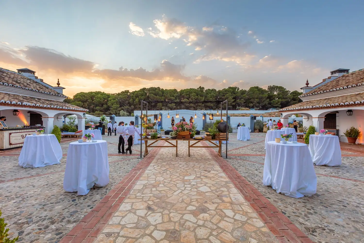 El patio empedrado al atardecer, con mesas altas de cóctel con mantel blanco bajo los arcos de la casa