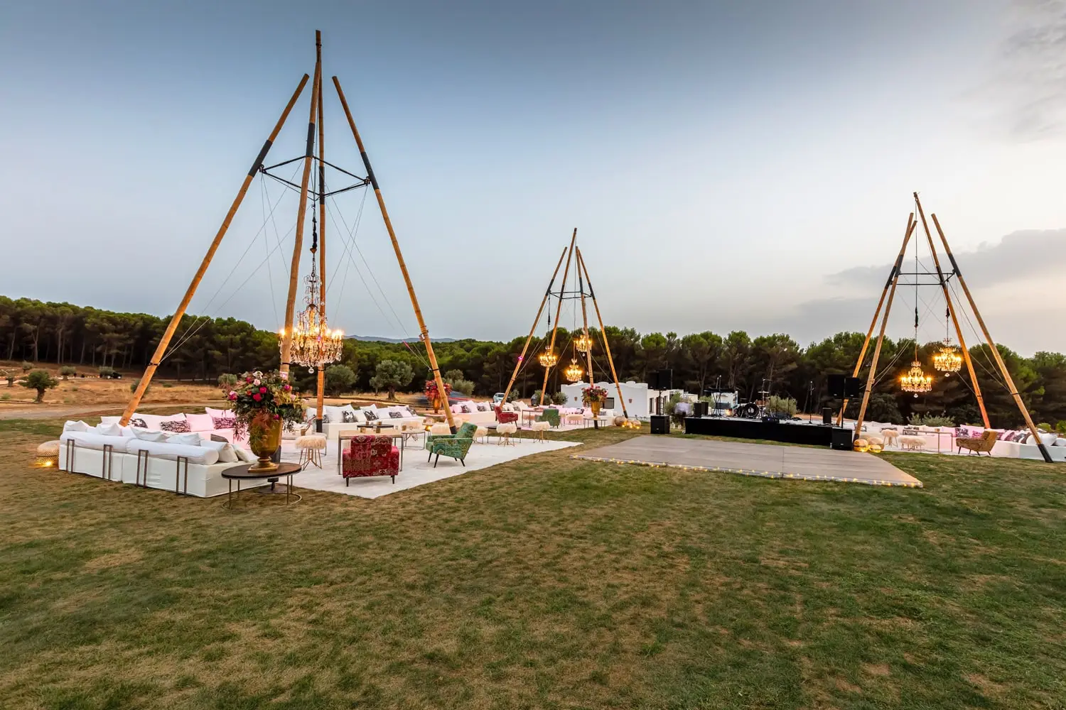 Three timber tripods with chandeliers above a lounge of sofas on the meadow, with the dance floor and stage beyond, at dusk